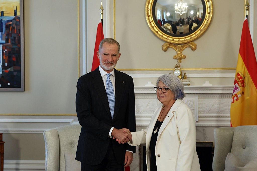 Two people standing in front of a fireplace shake hands, with flags of Spain and Canada behind them.