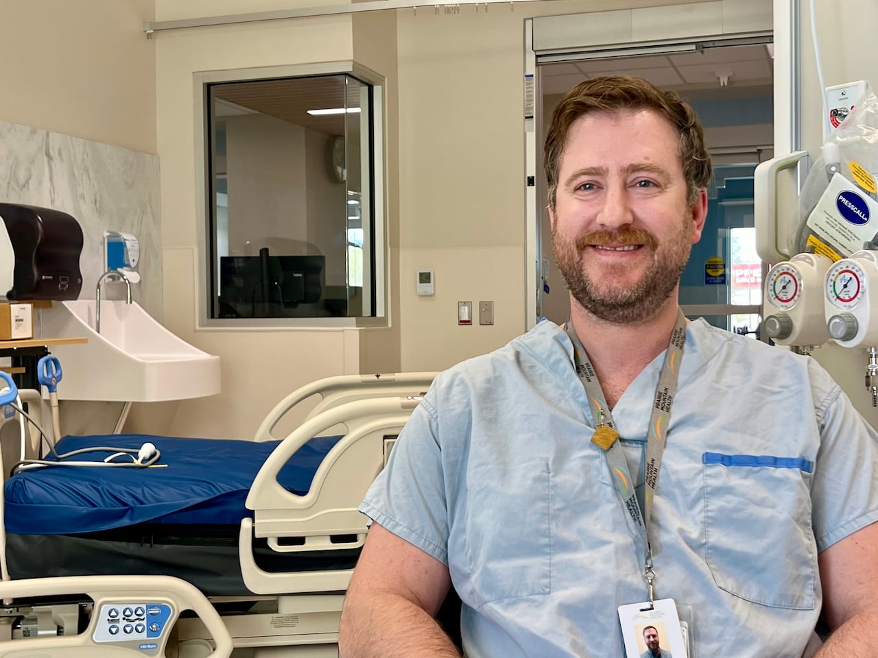 A man is smiling, sitting on a chair in a hospital room wearing light blue medical scrubs
