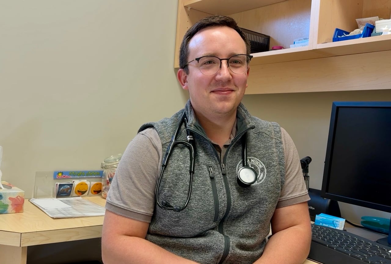 A man wearing a short-sleeve shirt and vest has a stethoscope around his neck and is sitting on a chair beside a blank computer screen