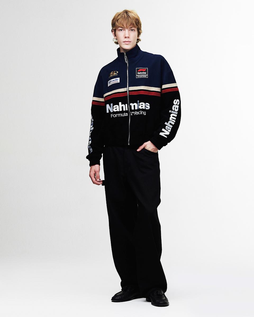 A person with blonde hair wearing a black "Nahmias" Formula 1 Racing jacket and black pants, standing against a plain background.