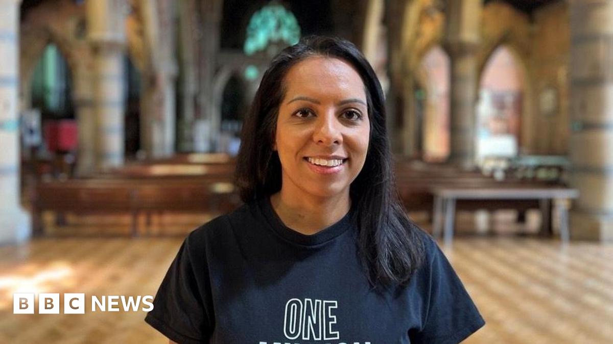 Amandeep is wearing a dark blue t-shirt, smiling at the front of a church