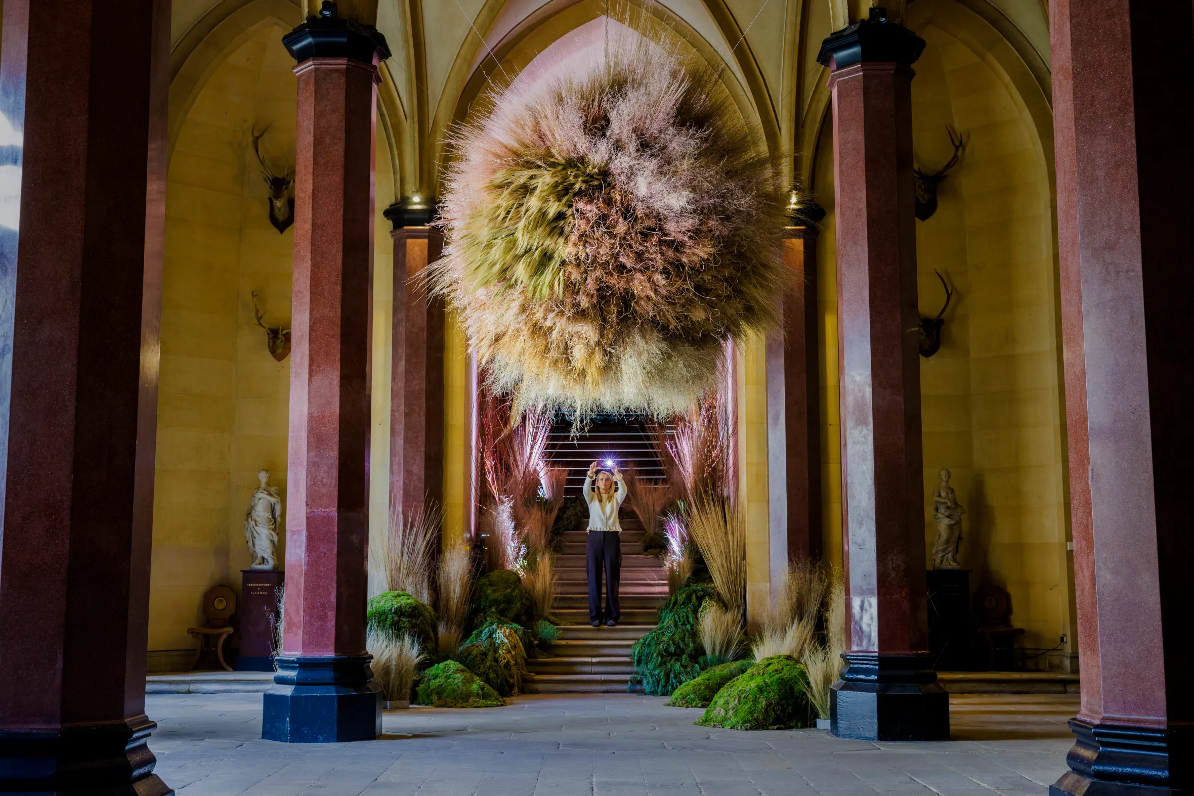 A visitor admires a suspended orb installation of dried deschampsia grasses in a castle hall.