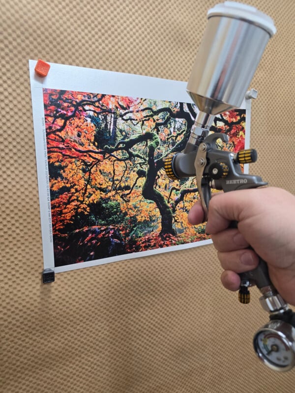 A hand holds a paint spray gun aimed at a colorful autumn tree photograph pinned to a textured brown surface with clips at the corners.