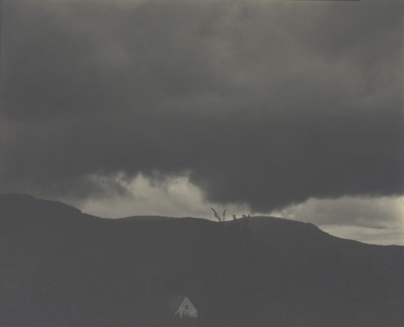A small white house sits beneath dark, dramatic storm clouds that loom over rolling hills, creating a moody and atmospheric landscape.