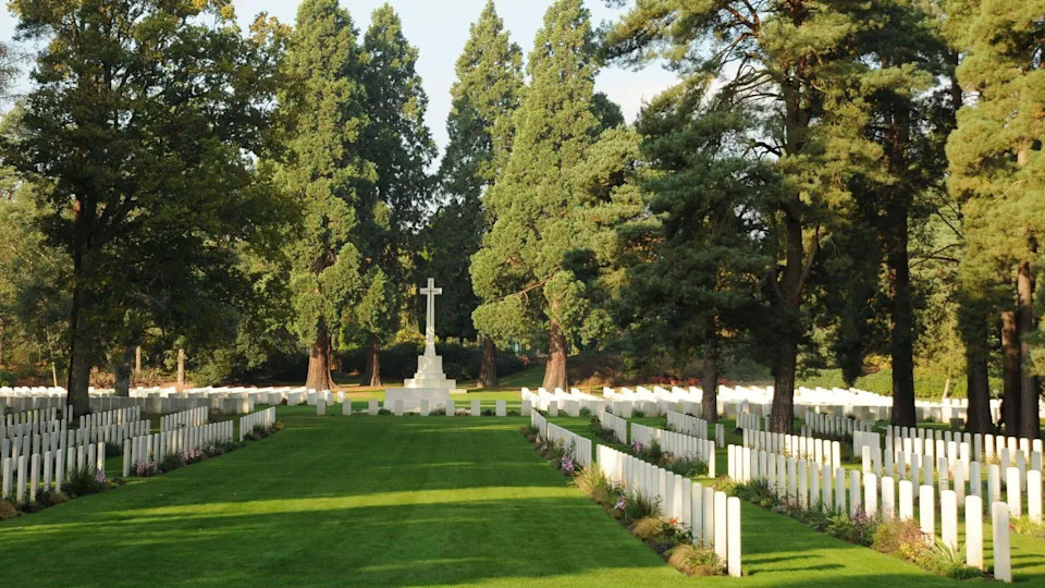 A shaded green lawn with rows of square white headstones, a large stone cross monument, and tall green trees behind.
