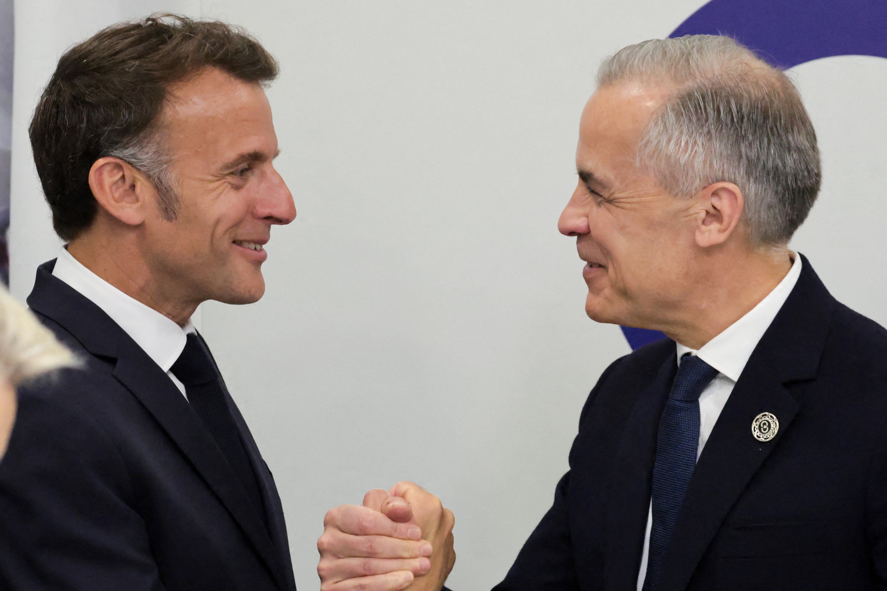 Emmanuel Macron and Mark Carney shake hands and smile at each other.