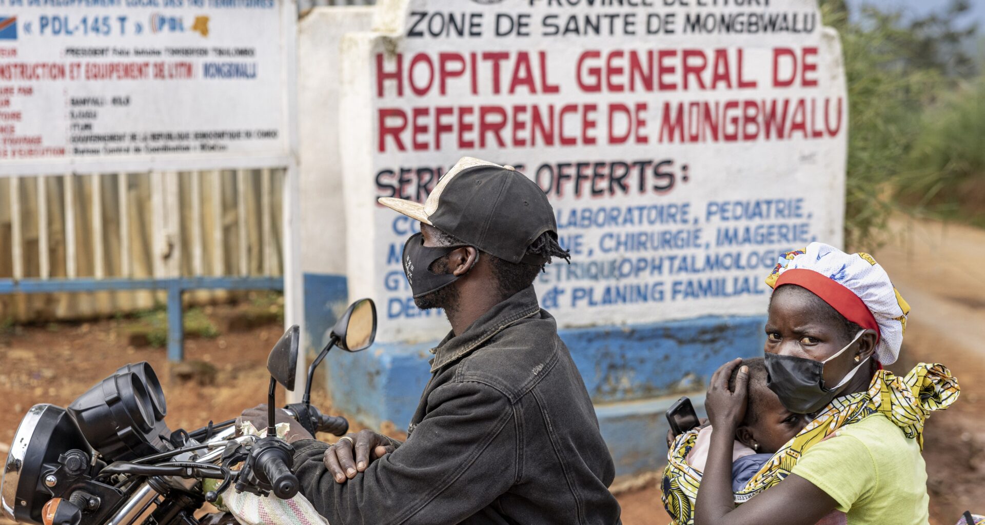 A woman holds a baby while riding on the back of a motorcycle. Behind them is a sign that announces the Hopital General de Reference de Mongbwalu