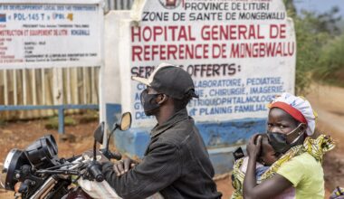 A woman holds a baby while riding on the back of a motorcycle. Behind them is a sign that announces the Hopital General de Reference de Mongbwalu