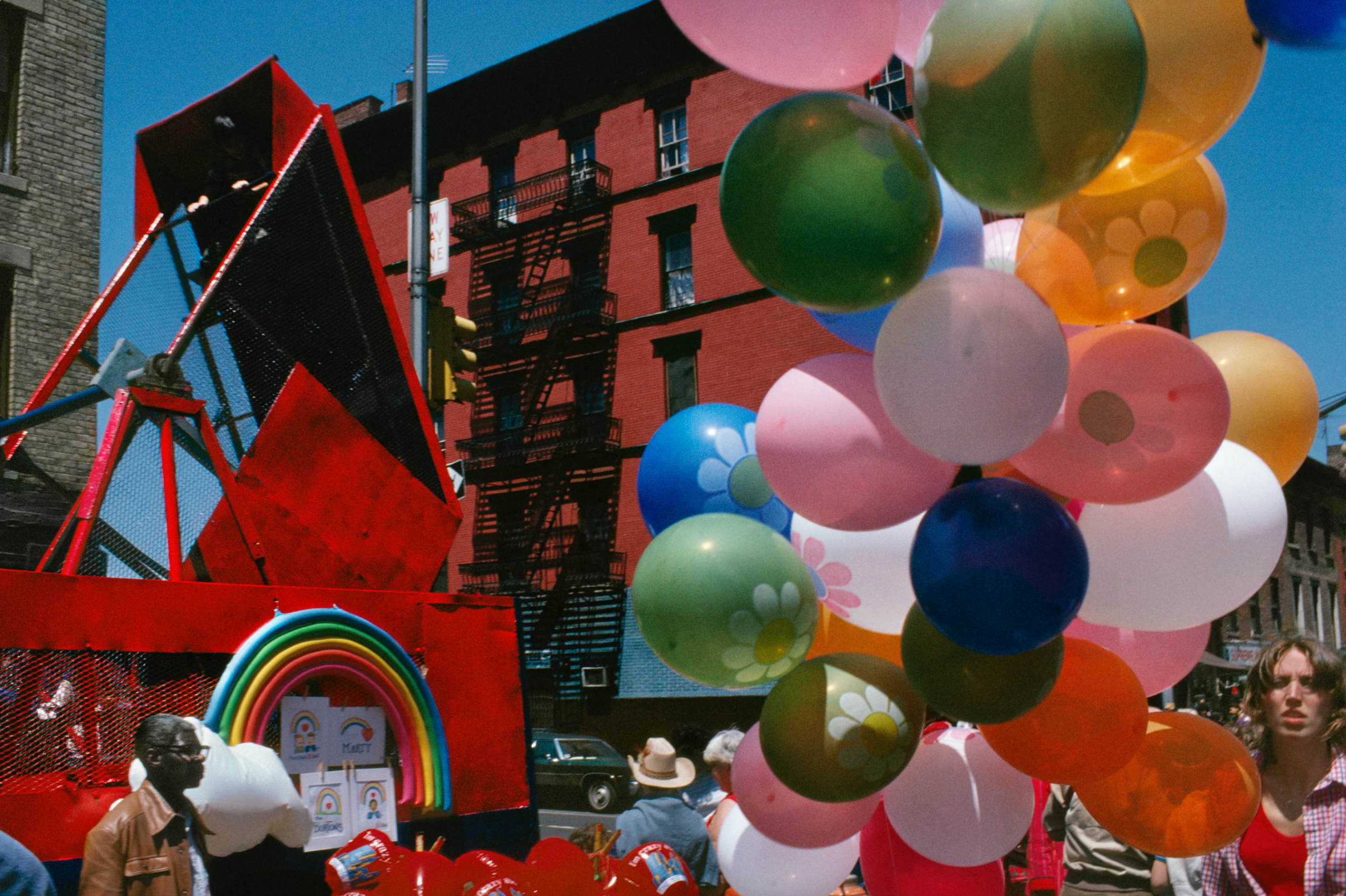 A street scene with many colorful balloons in the foreground, and a red building with a fire escape in the background.