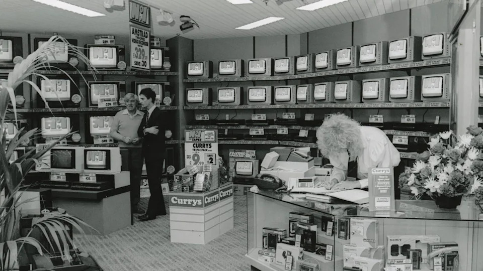 A black and white photo of the technology shop Currys in the 80s. There is a blonde woman with a perm bent over behind the counter. Two men talk in the left corner of the shop. The back wall is lined with old, small televisions. There is a plinth with Currys written on it in the centre of the photo.