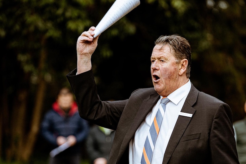 An auctioneer shouts with a paper rolled up in his raised hand.