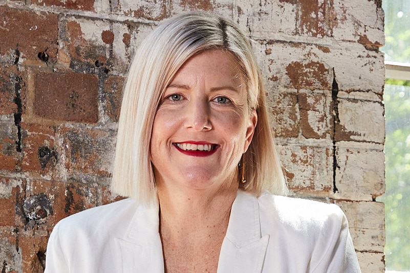 A headshot of a smiling woman with a blond bob, wearing a white blazer and shirt.
