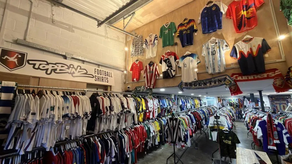 A shop interior with hundreds of football shirts of different colours. The name of the shop "TopBinz Football Shirts" is displayed on the wall next to their red, white and black logo.