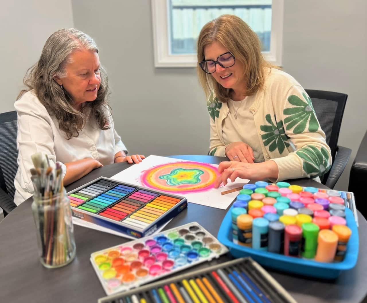 Two women sit at a round table with an artwork, surrounded by paints and crayons.