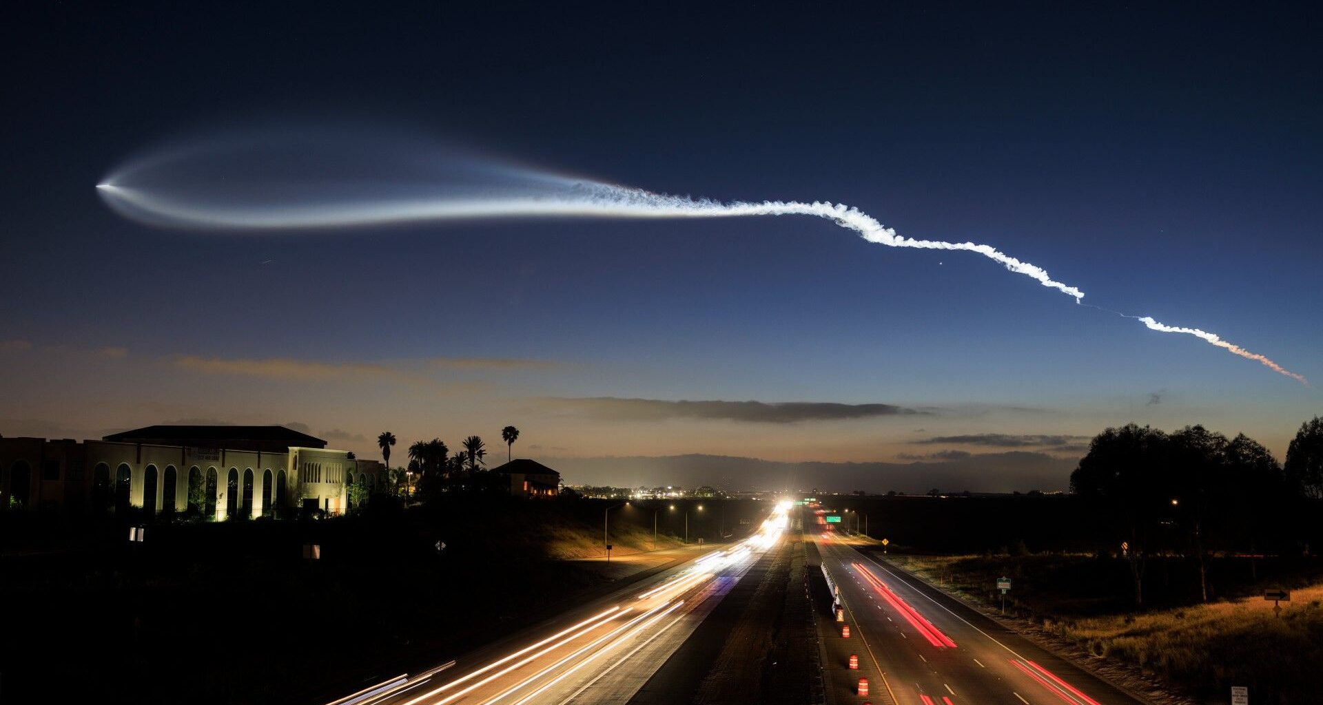 a streak of white gas stretches across a nearly dark twilight sky above a freeway lined with palm trees