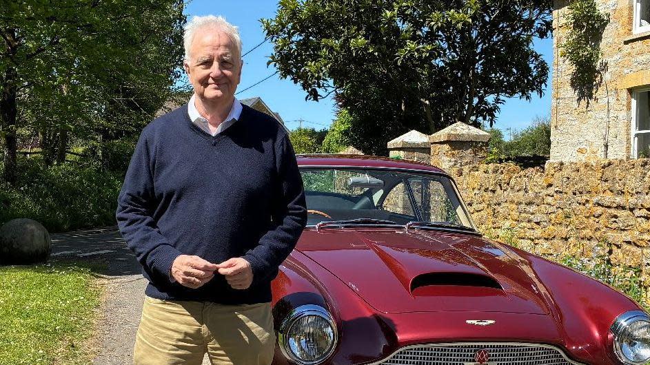David Garmston stands in front of a deep red Aston Martin DB4. He has short white hair and is wearing a blue shirt underneath a navy-blue jumper and sand-brown chinos. The picture has been taken on a sunny day and there is a cottage in the background.