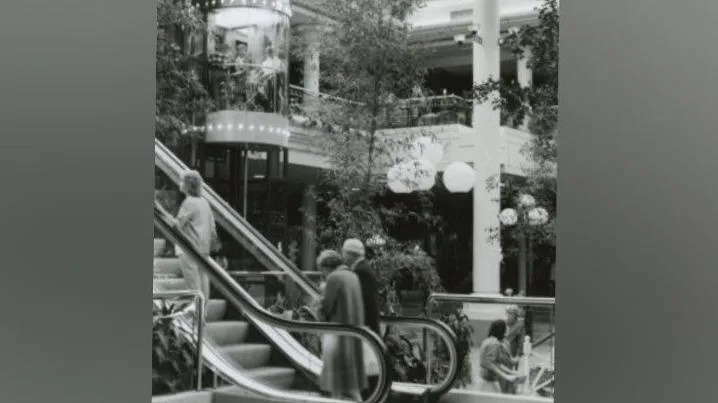 A black and white photo of an older couple going up on an escalator. There is a woman in front of them. To their right there is a tubular lift which has people inside of it. There is a white pillar in the centre of the room which is large and open with a glass ceiling. There are trees decorating the space.