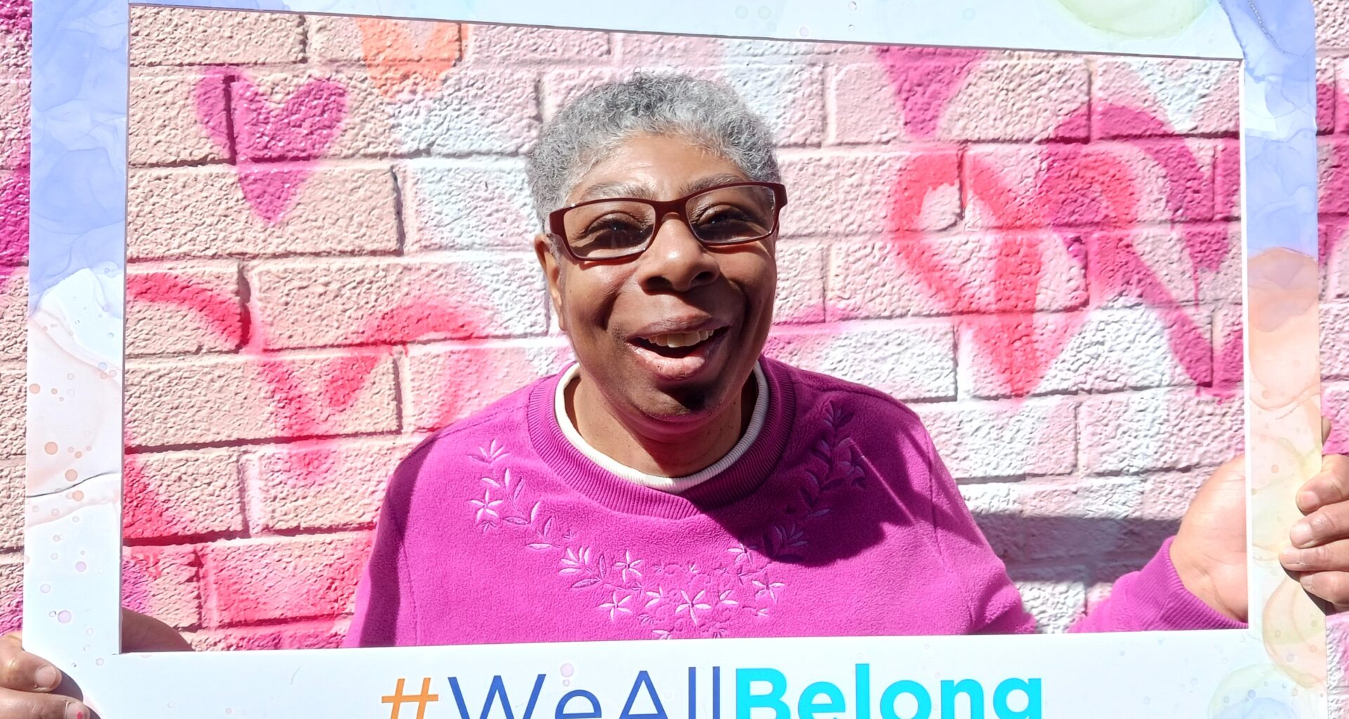 Photo of a smiling woman in a pink sweater holding a #WeAllBelong frame.