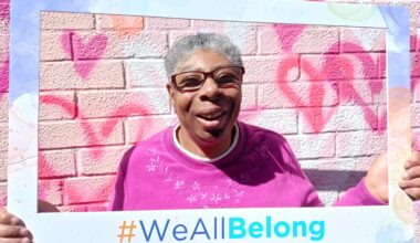 Photo of a smiling woman in a pink sweater holding a #WeAllBelong frame.
