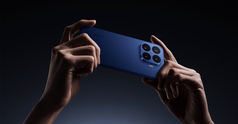 Two hands hold a blue smartphone horizontally against a dark background, showing the large quad-camera module on the back and the Xiaomi logo.