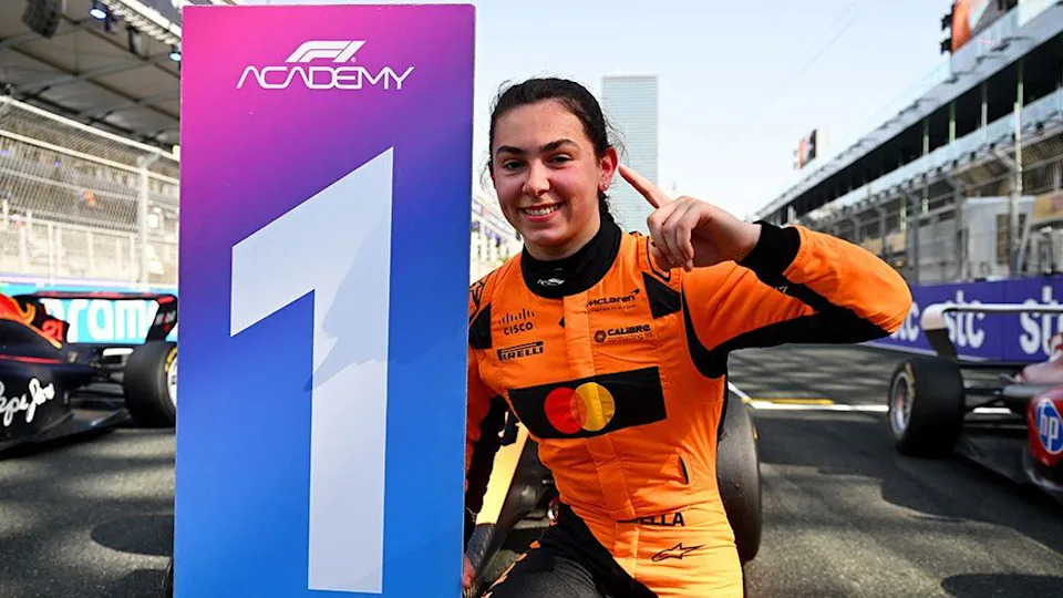 Ella Lloyd, wearing an orange race suit with black sponsor logos, holding up a "one" sign with her fingers while crouching next to a "one" winners board. She is on the race track in front of a Formula 1 car.