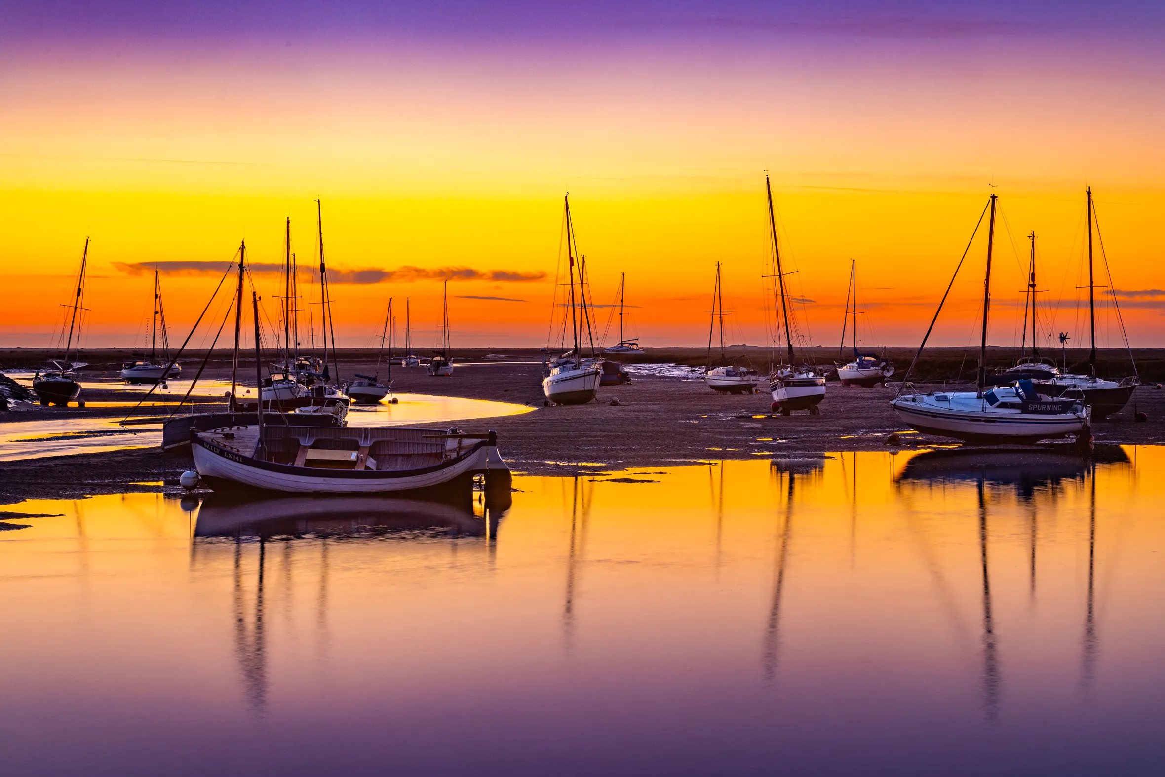 Sailboats moored in Wells-next-the-Sea harbour at dawn, with a vibrant orange and purple sky reflecting on the water.