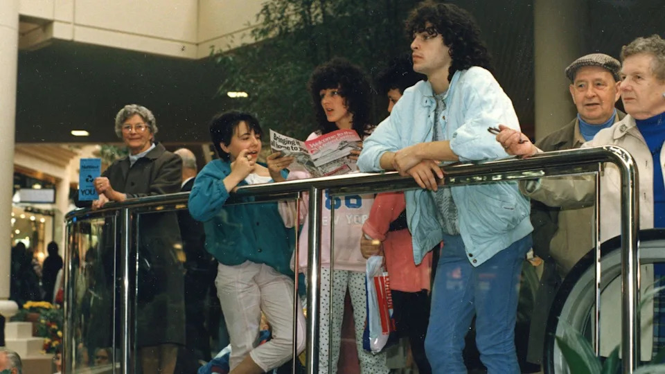 A group of people standing behind a glass barrier. There is an older lady with short, grey hair on the left looking at the camera while holding a blue leaflet. Next to her are three young women with dark hair, wearing 80s-looking clothes. Next to them on the right is a young man with curly dark hair wearing a baby blue bomber jacket. He is leaning over the railing. There is an older couple on the far right looking out in front of them. Could be in the 1980s.