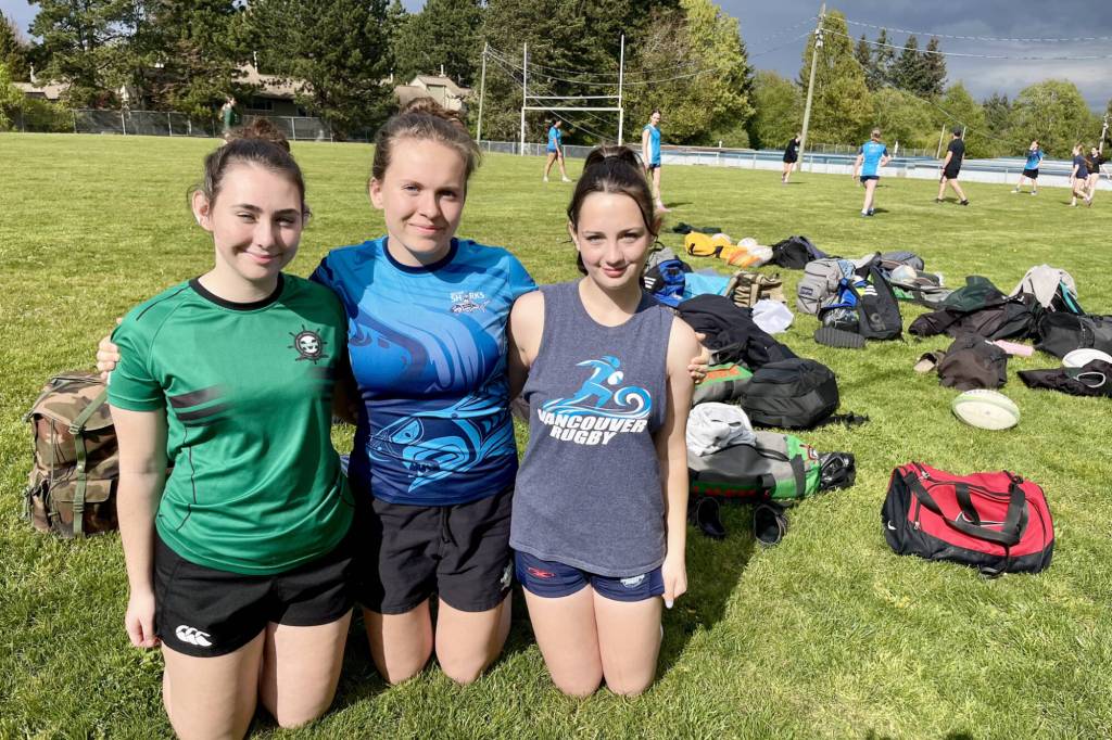 Grade 12 students Cooper Marsden, left, Astrid Maynes and Isla Van Staveren pose for a photo as their Earl Marriott Secondary girls rugby team starts practice in the background. The Mariners squad has started out their season 3-0 so far. (Tricia Leslie photo/Peace Arch News)