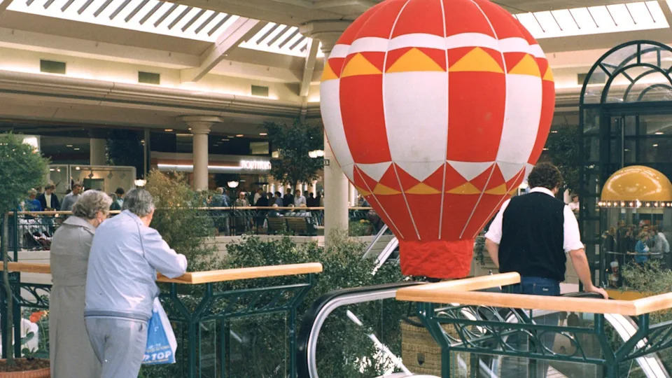The upper balcony of the shopping centre with people leaning over the wooden bannister. In the open space in the centre there is a small inflatable red, white and yellow hot air balloon.