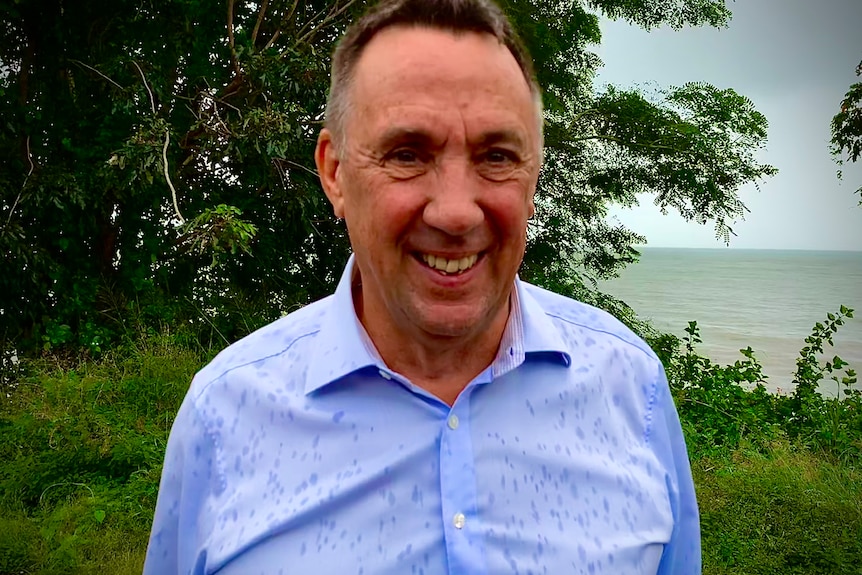 Portrait of man in blue shirt by sea with trees behind.