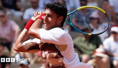 Adolfo Daniel Vallejo, wearing a white patterned t-shirt, plays a backhand with his yellow, blue and black racquet held in two hands over his right shoulder