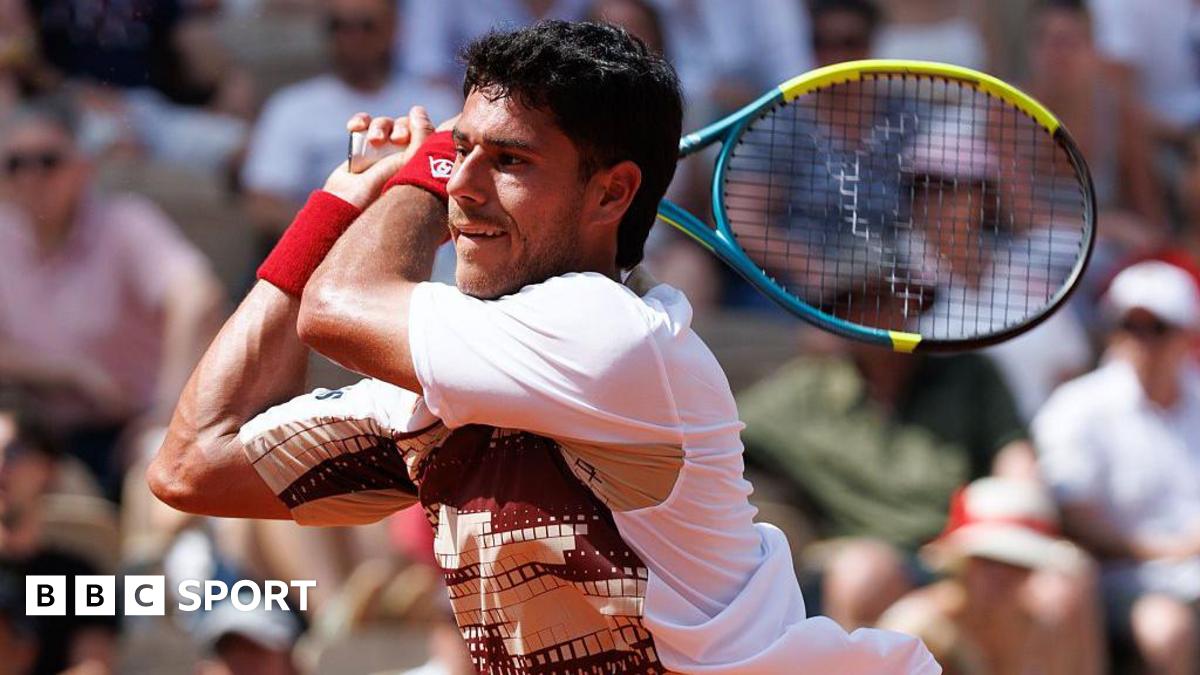 Adolfo Daniel Vallejo, wearing a white patterned t-shirt, plays a backhand with his yellow, blue and black racquet held in two hands over his right shoulder
