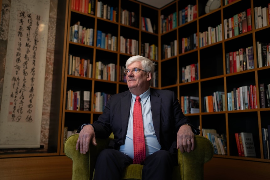 A man with glasses and a suit and tie sits in a light green chair surrounded by books in his personal library.