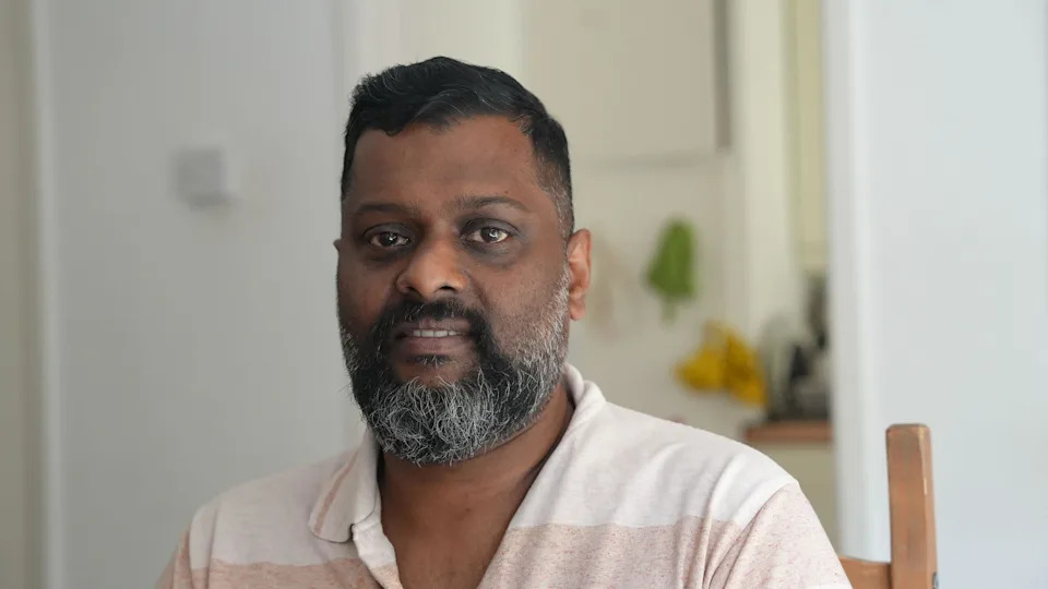 Sumesh has a dark beard and is wearing a beige polo shirt. He is sitting in a wooden chair and smiling at the camera