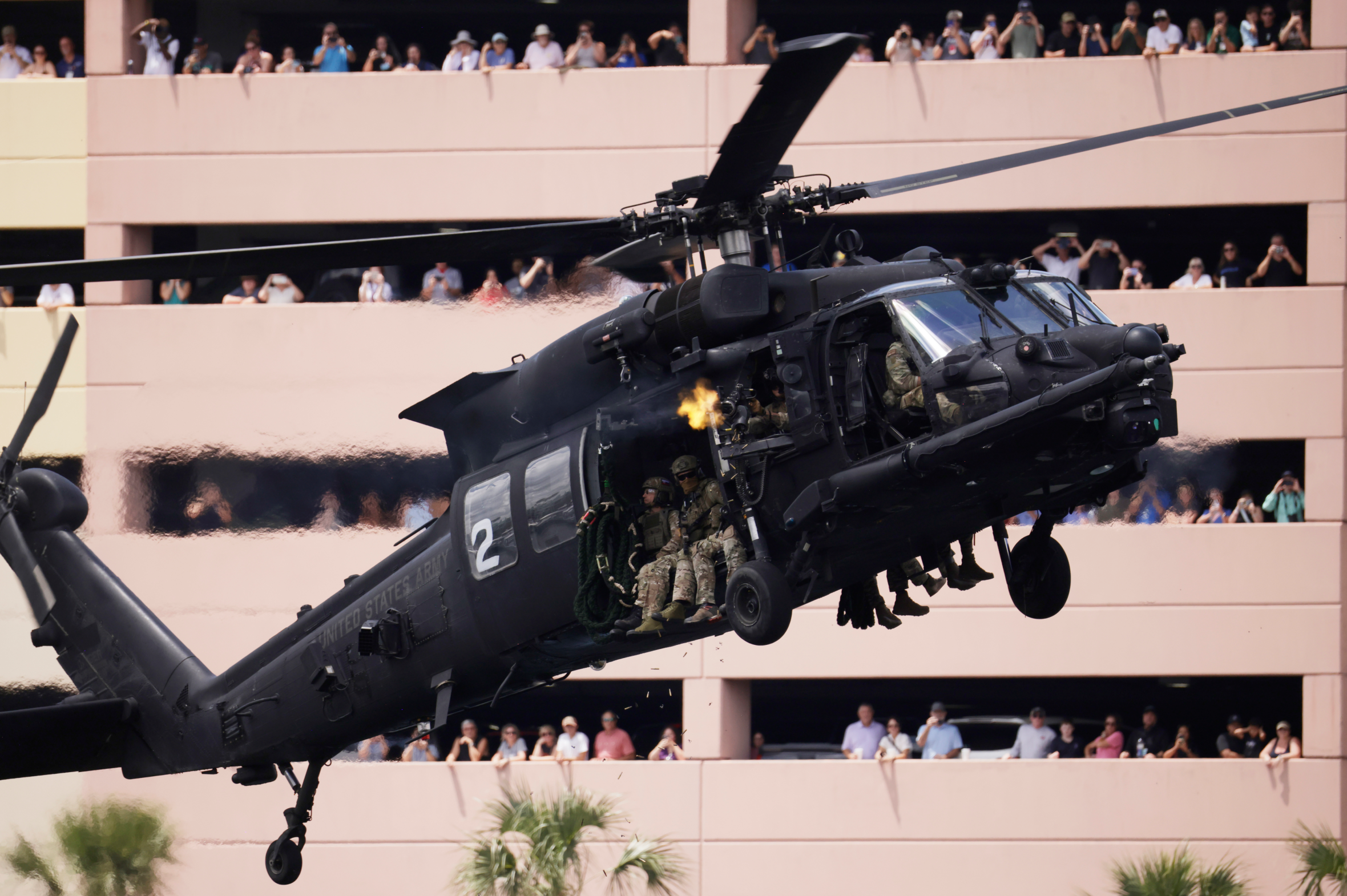 A black MH-60 Blackhawk helicopter with military personnel inside, one firing a weapon with flames and smoke, flies in front of a building where spectators watch from balconies.