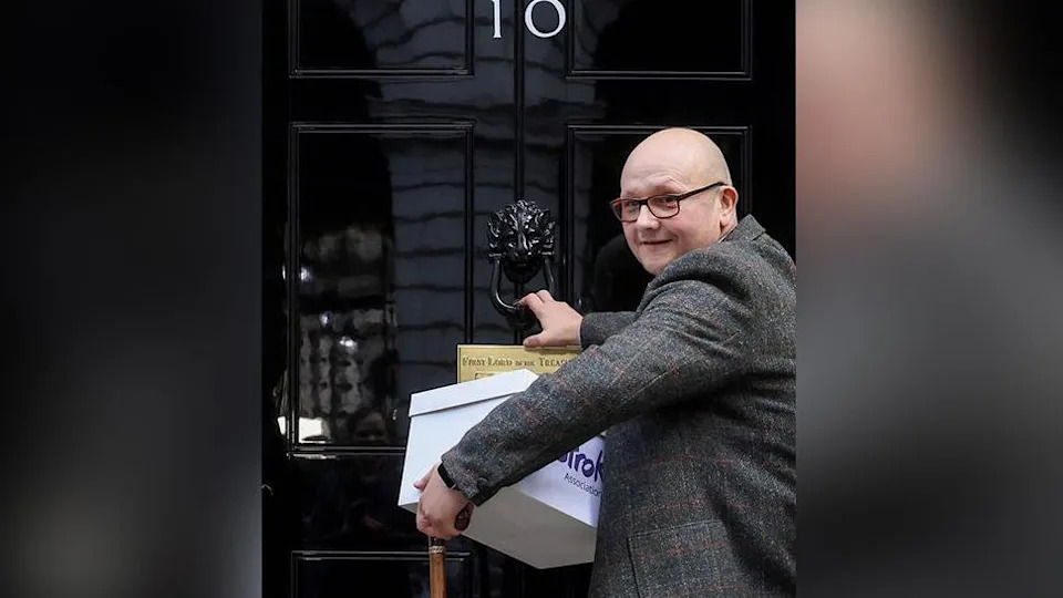 Campaigner Phil Woodford who is bald and wearing glasses and a tweed blazer knocks on the door of No 10 Downing Street with a box of signatures. He is smiling.