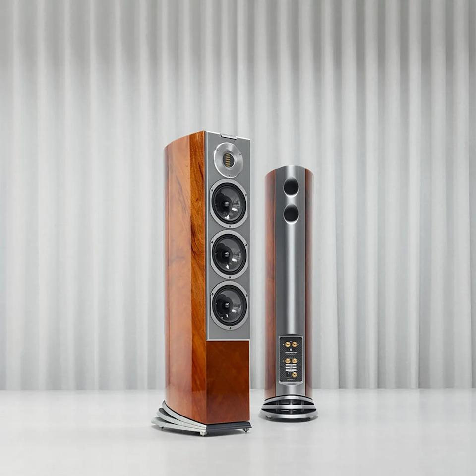 Pair of tall floor-standing speakers with wood veneer sides, silver front panels, and multiple drivers, set against a gray curtain backdrop.
