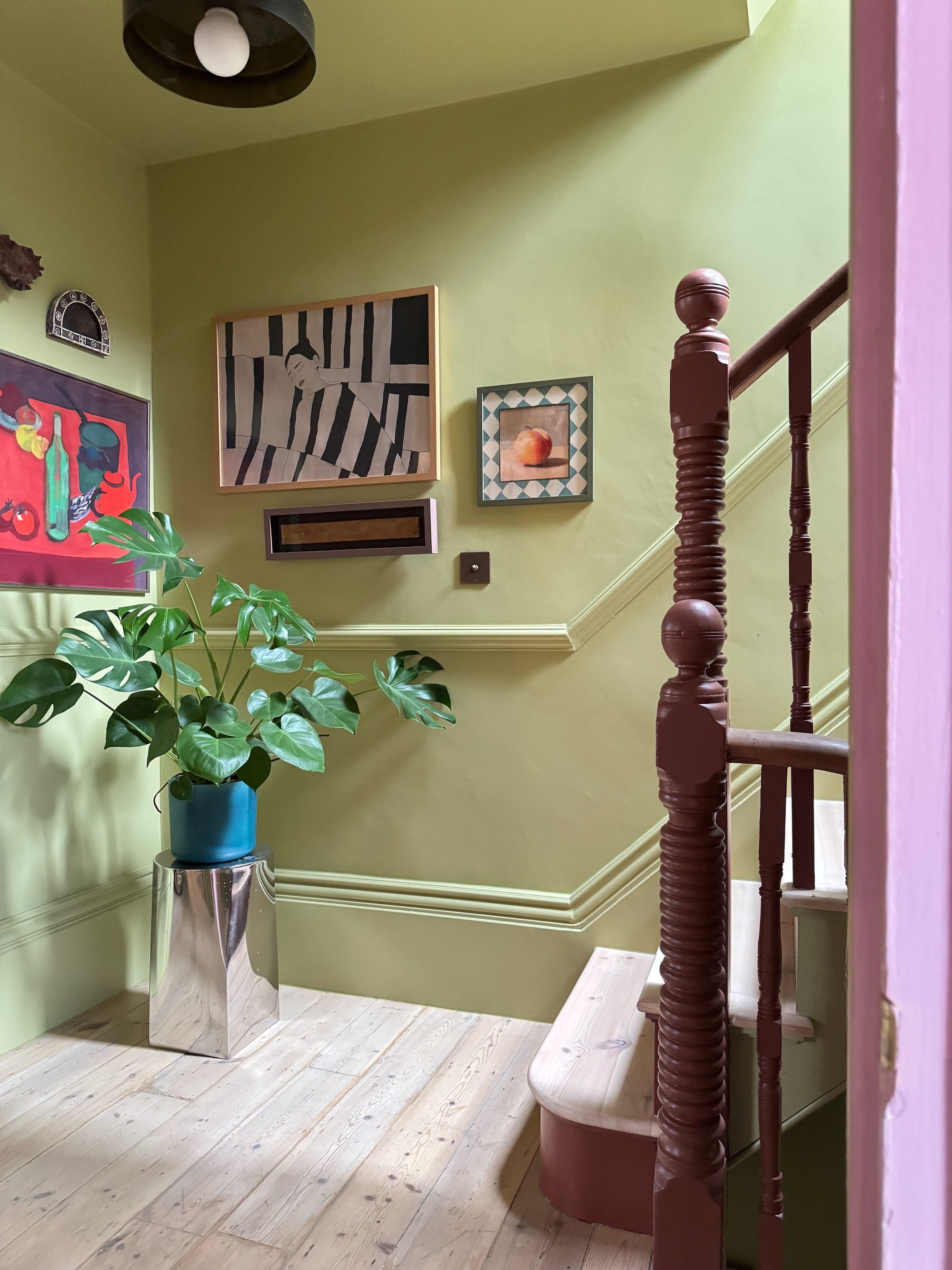 Image of a hallway/stairwell with powdery, lime green walls. There is a gallery wall of modern art and a green plant on a chrome plant stand.