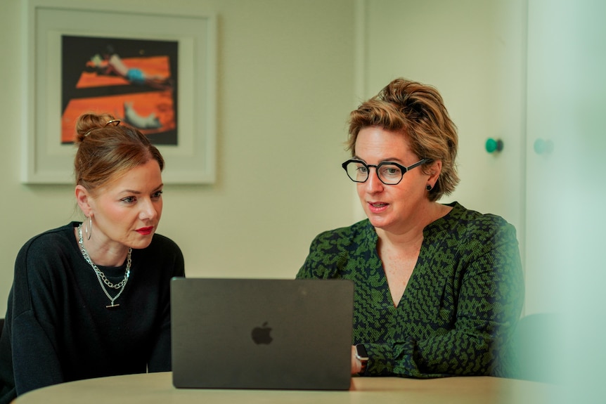 Annie on the left sits next to Angela at a table both looking at a laptop