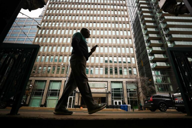 A silhouette of a person walking while using a cell phone. They are in front of a tall, grey office building with small glass windows in downtown Toronto as car passes in the street behind.
