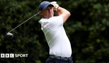 Rory McIlroy at the Truist Championship, wearing a white golf shirt and blue cap, swinging a metal wood