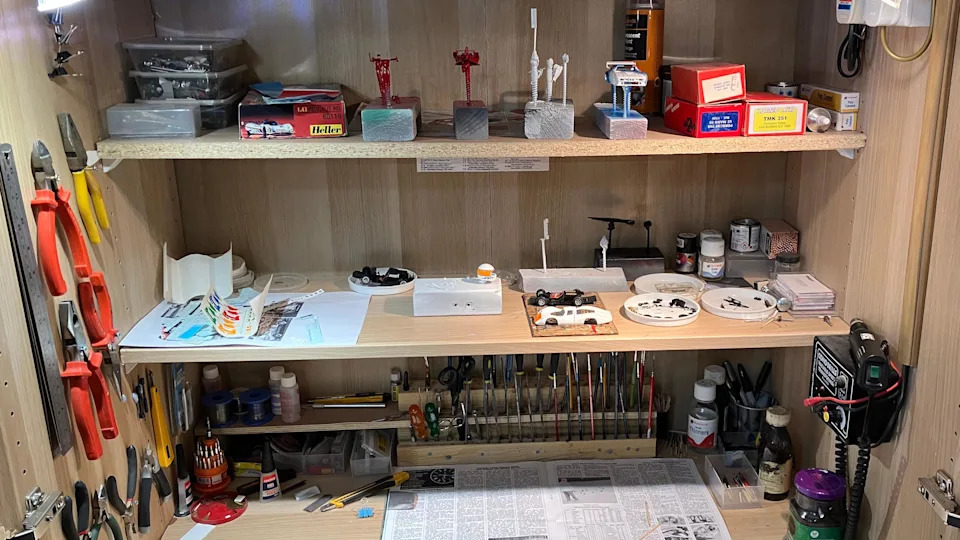 A compact workshop shelf is filled with model‑making tools, paints, brushes, and small parts neatly arranged.
Miniature car bodies, stands, and boxed kits sit alongside clamps, pliers, and precision instruments.
The setup suggests a dedicated, well‑used space for detailed scale model construction and finishing.