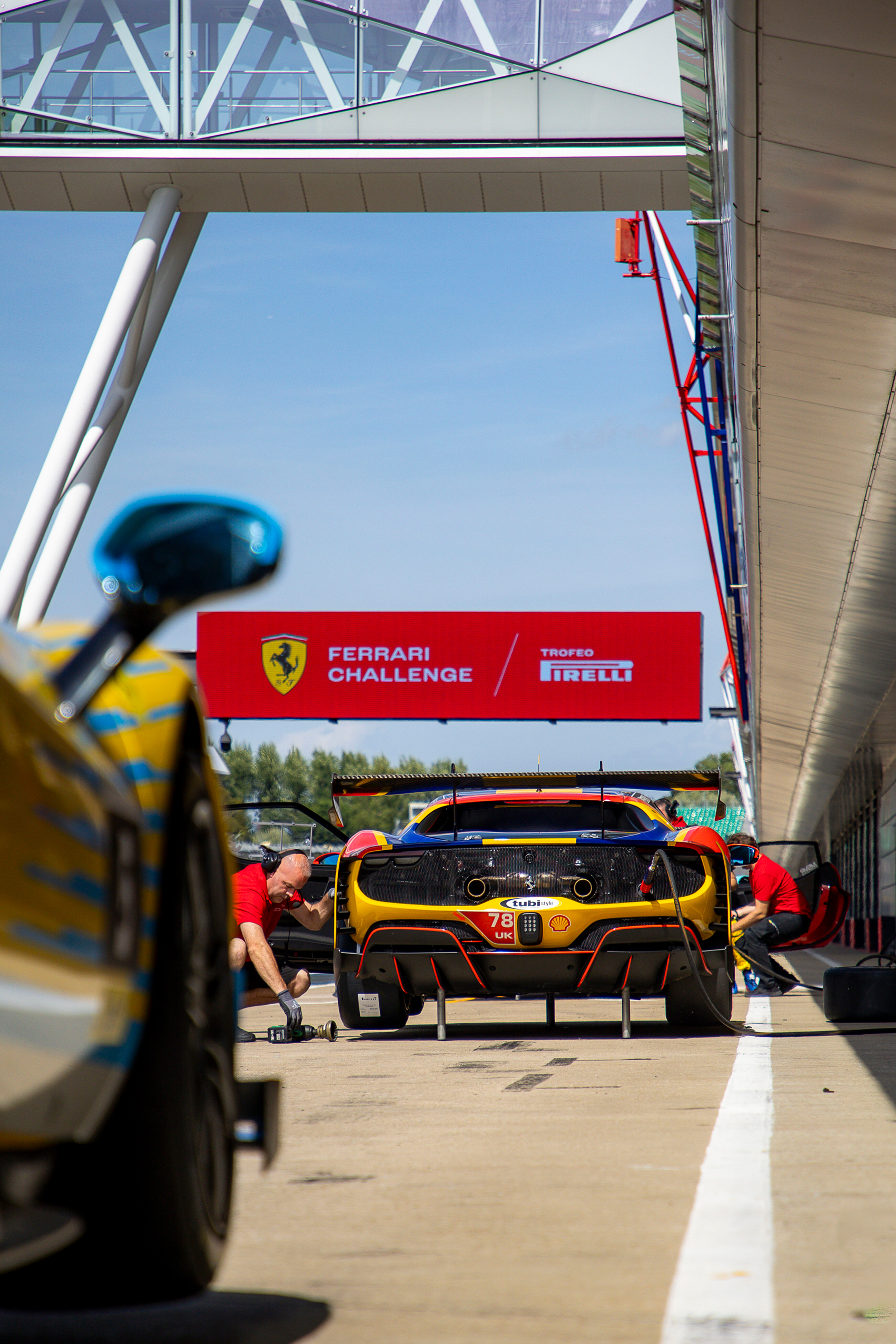Ferrari race car in pit lane with mechanics working under a clear blue sky, Ferrari Challenge and Pirelli sign in background