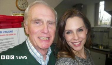 Don Taylor, who has white, receding hair and is wearing a green jumper and a white and blue check shirt, and and his daughter Emma Kenny, who has long brown hair and is wearing a grey top, are posing for a selfie in a kitchen. They are both smiling at the camera.