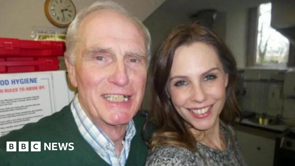 Don Taylor, who has white, receding hair and is wearing a green jumper and a white and blue check shirt, and and his daughter Emma Kenny, who has long brown hair and is wearing a grey top, are posing for a selfie in a kitchen. They are both smiling at the camera.