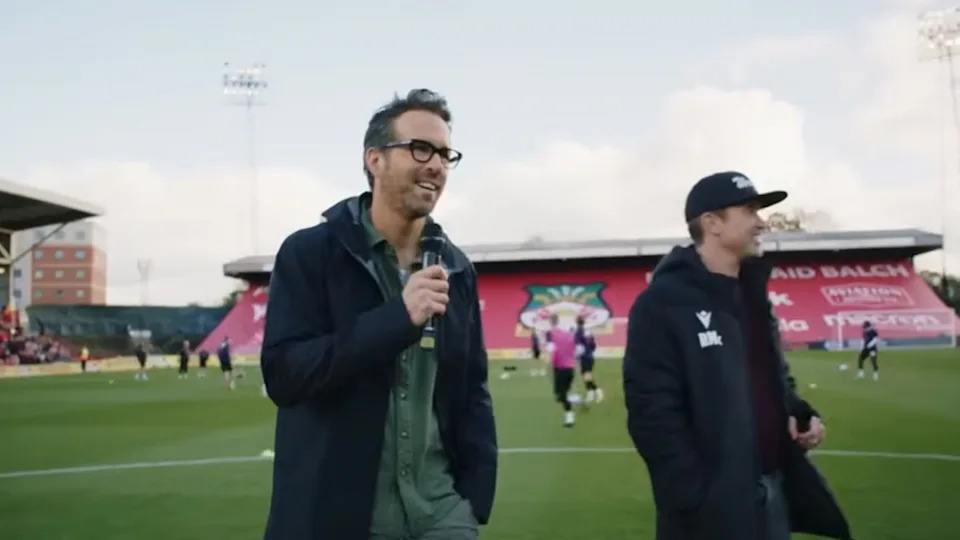 Ryan Reynolds talking into a microphone on a football pitch with Rob Mc Elhenney standing behind him in Welcome to Wrexham.