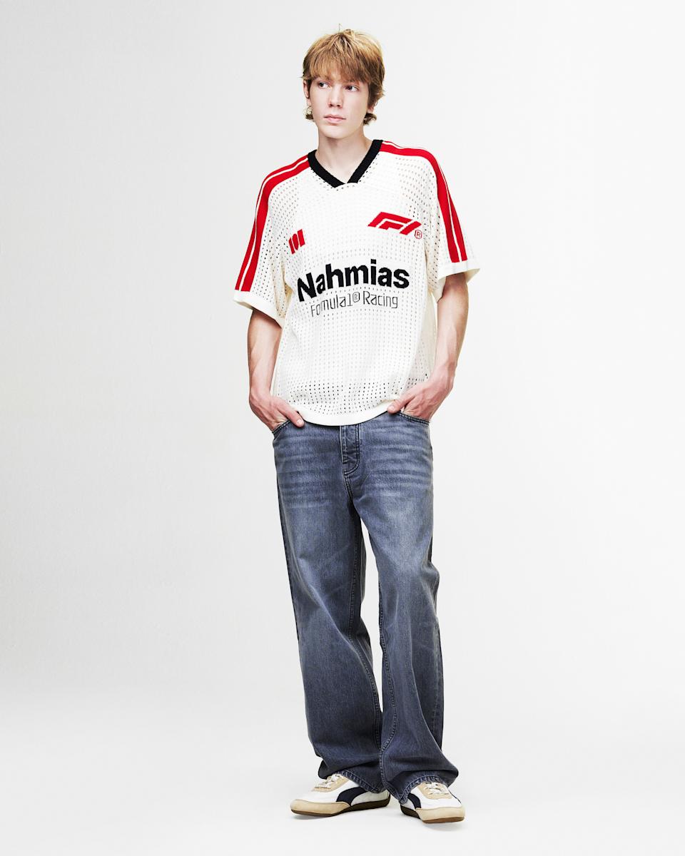 A person with blonde hair wearing a white mesh jersey with red stripes, blue jeans, and sneakers, standing against a plain background.