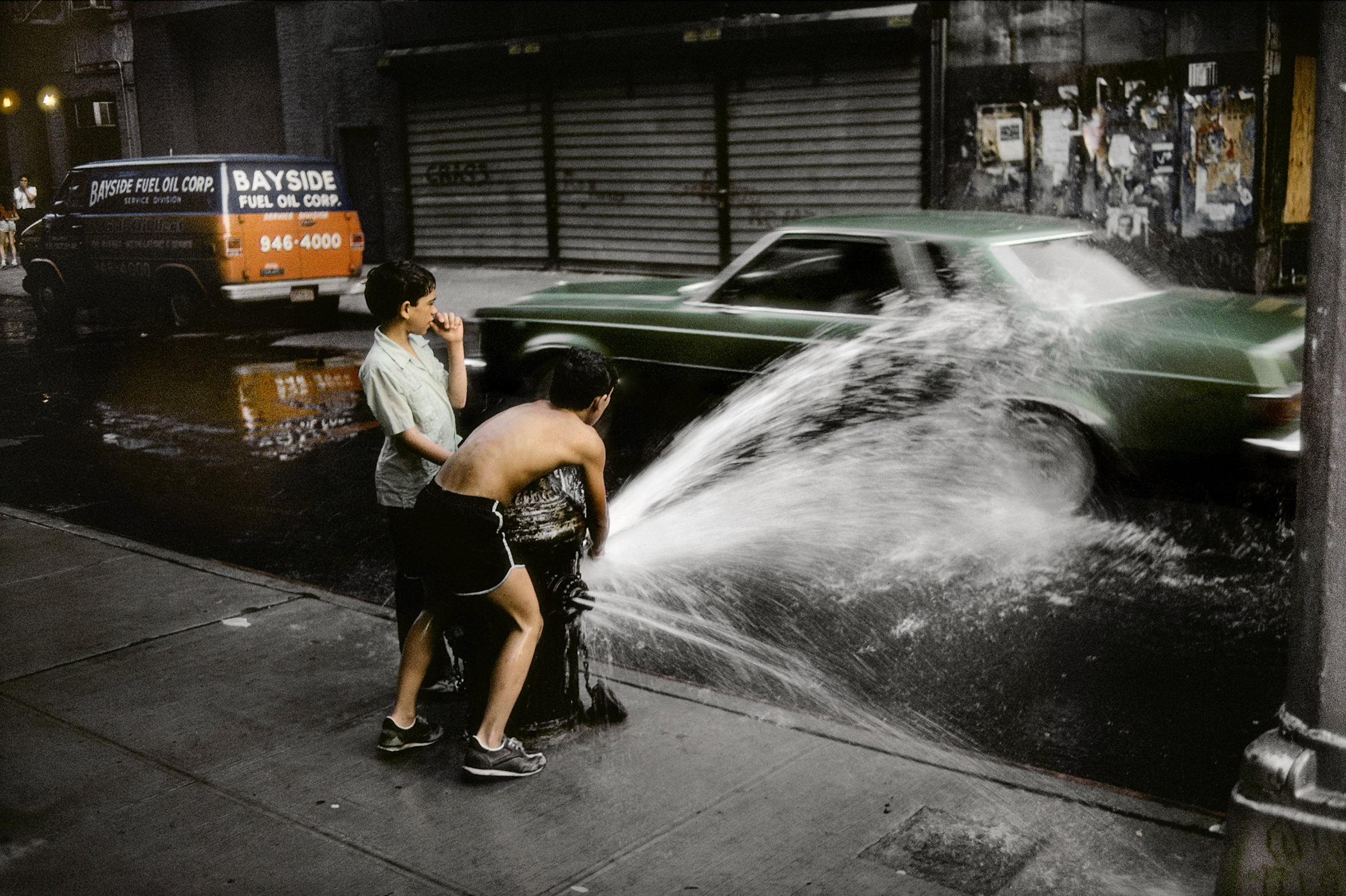 Two boys opening a fire hydrant on a city street, soaking a passing green car.
