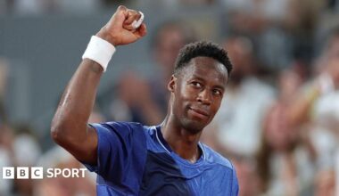 Novak Djokovic, in a pale yellow T-shirt, stands facing Gael Monfils, wearing a white T-shirt, rests his right arm on the Frenchman's shoulder