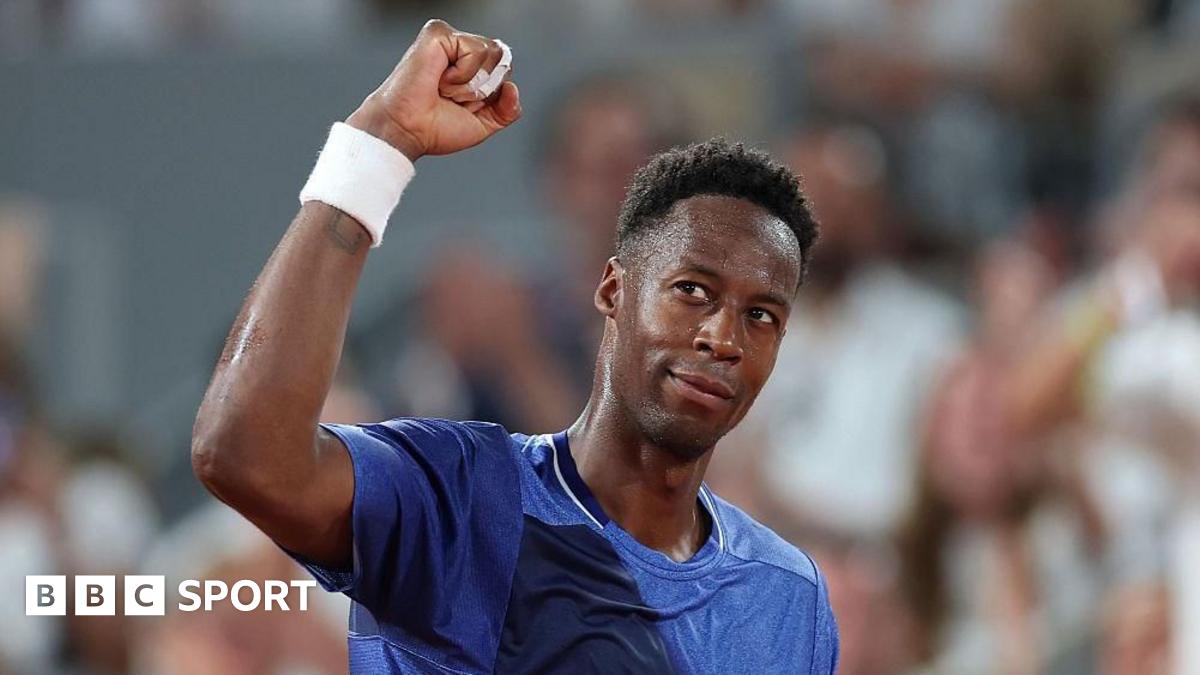 Novak Djokovic, in a pale yellow T-shirt, stands facing Gael Monfils, wearing a white T-shirt, rests his right arm on the Frenchman's shoulder